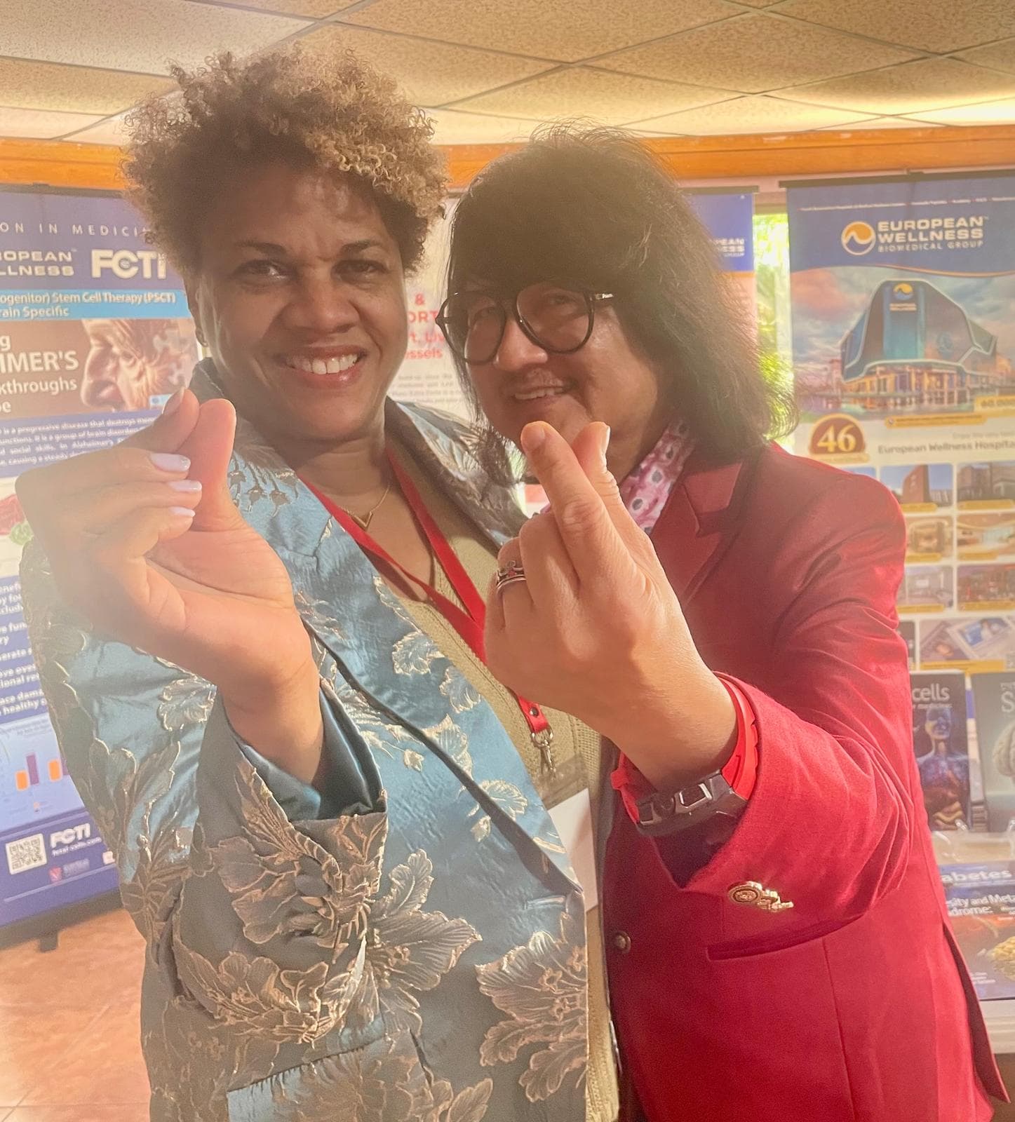 Smiling woman in blue floral blazer and man in red blazer making finger heart gestures.