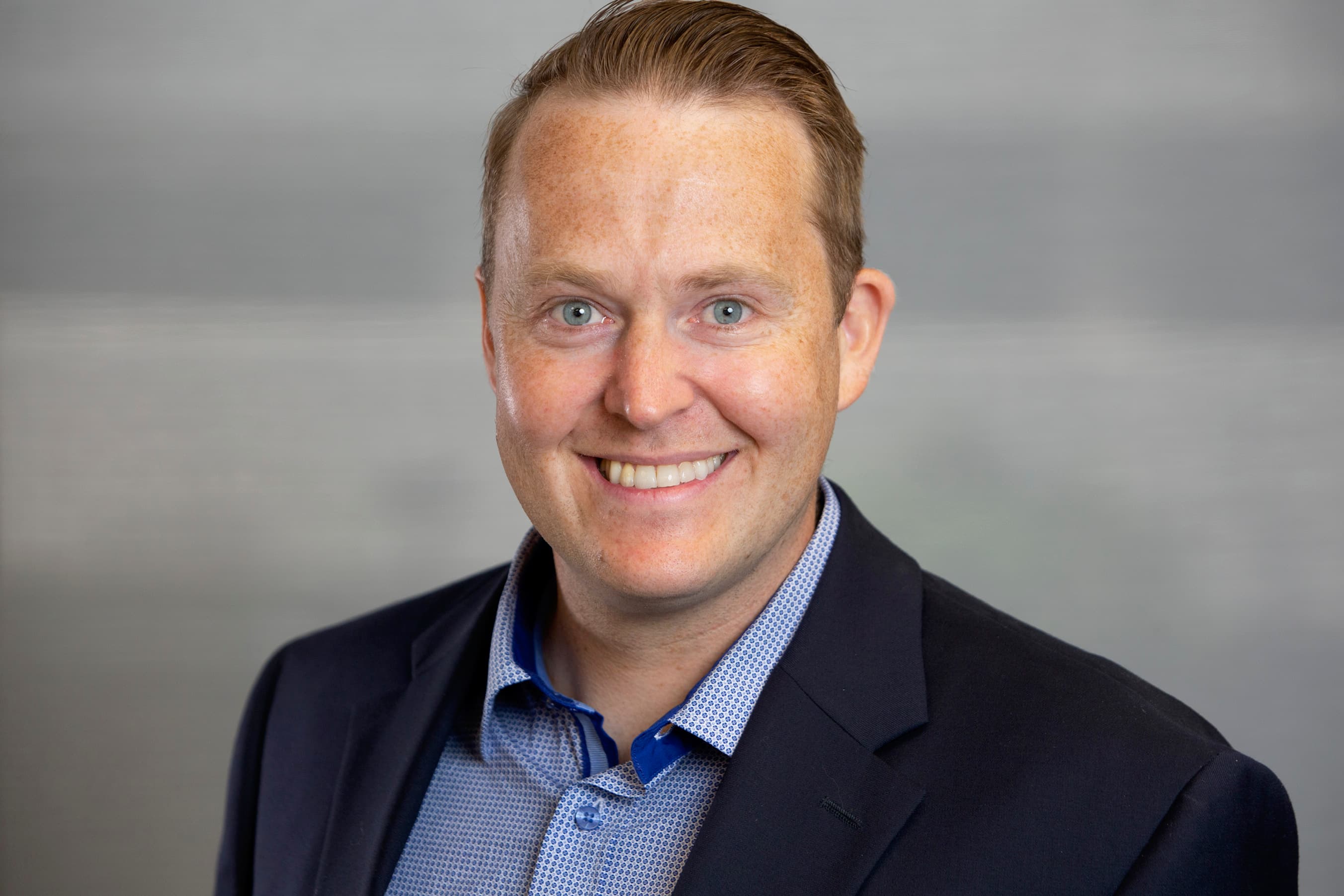 Smiling man with light brown hair and blue eyes wearing navy blazer and patterned shirt.