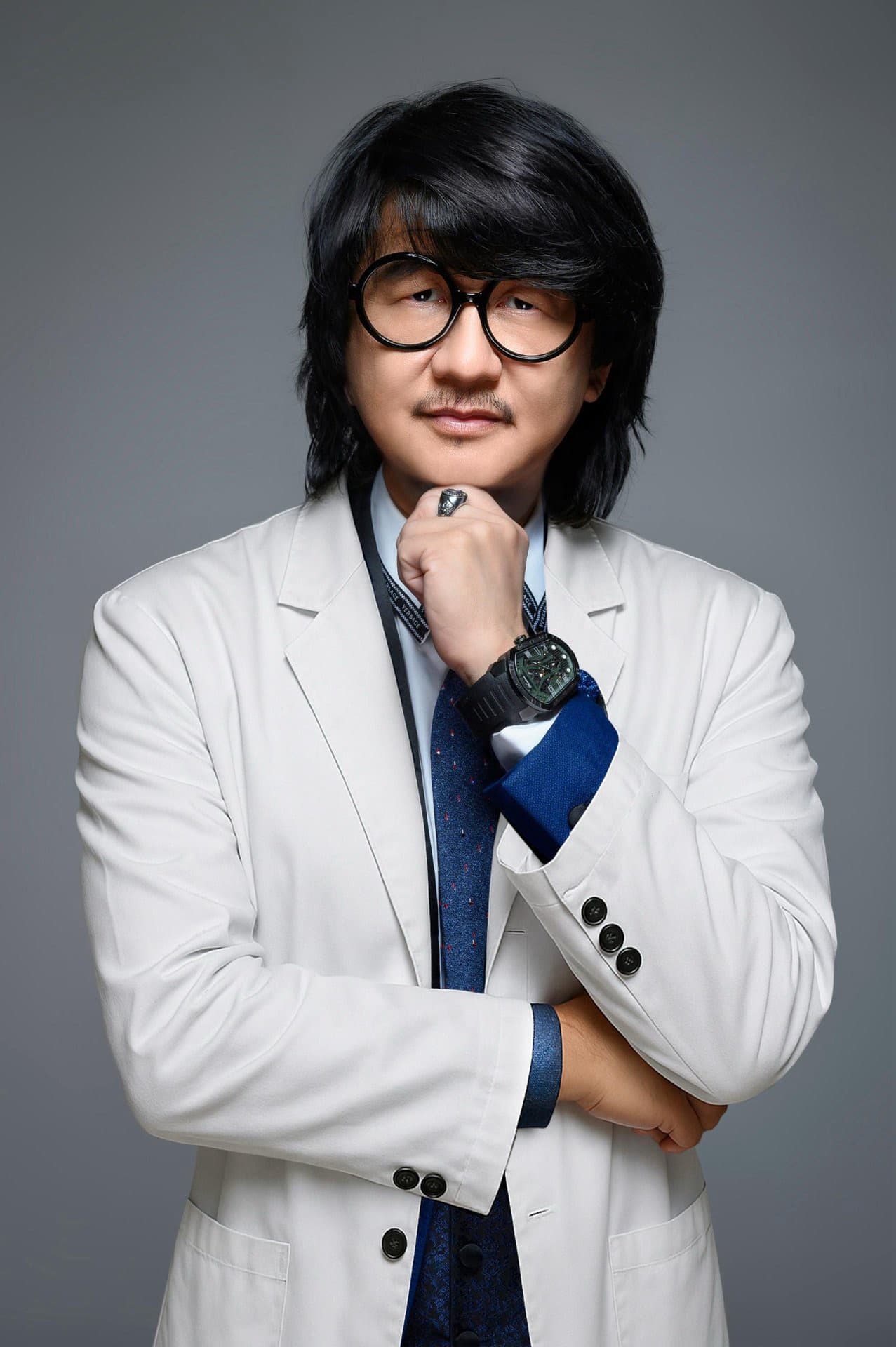 Man with round glasses and black hair wearing a white blazer and blue tie.