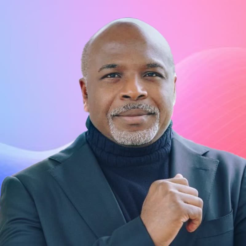 Smiling bald Black man with a grey goatee wearing a dark turtleneck and blazer.