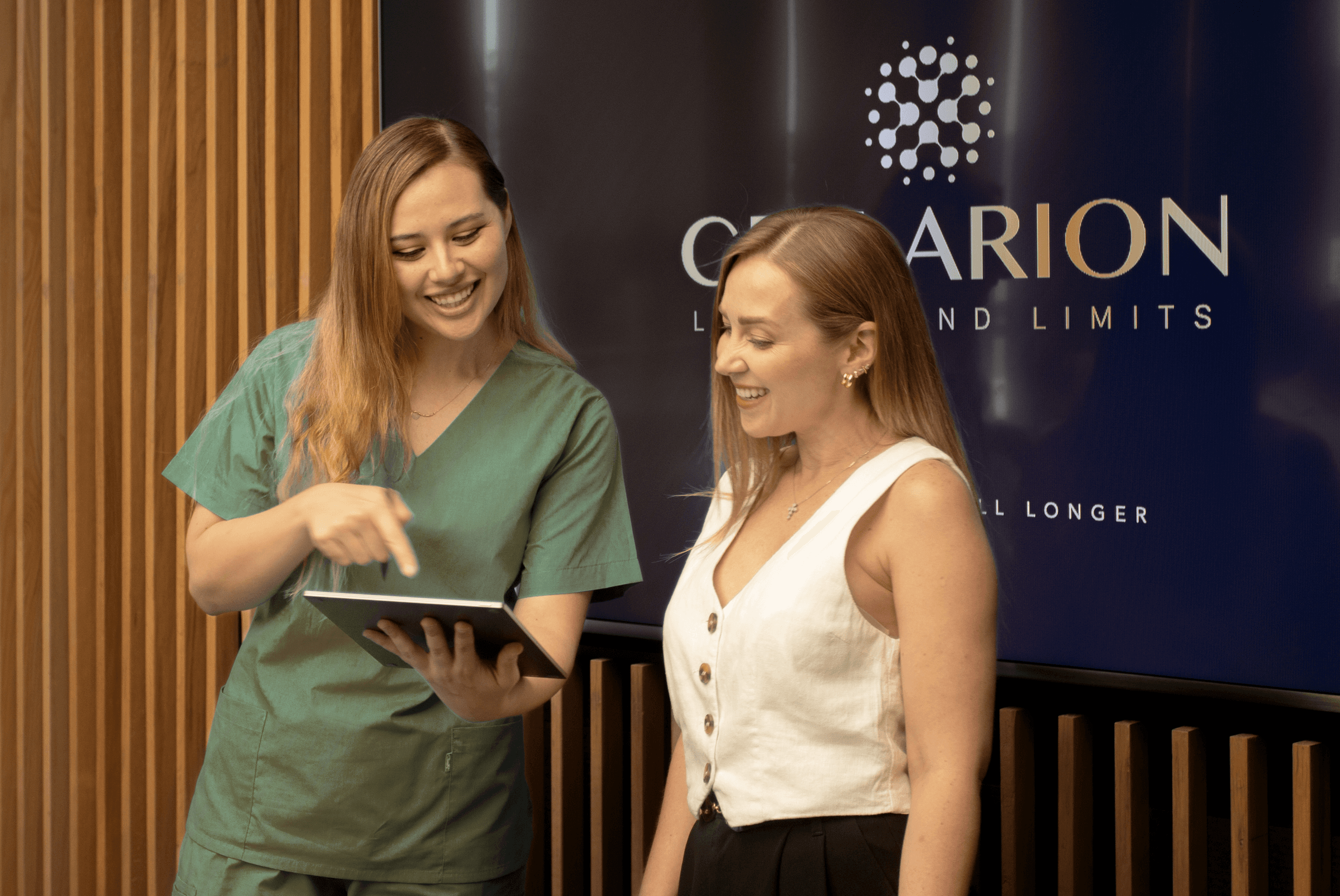Healthcare professional in green scrubs shows a tablet to a smiling woman in a clinic.